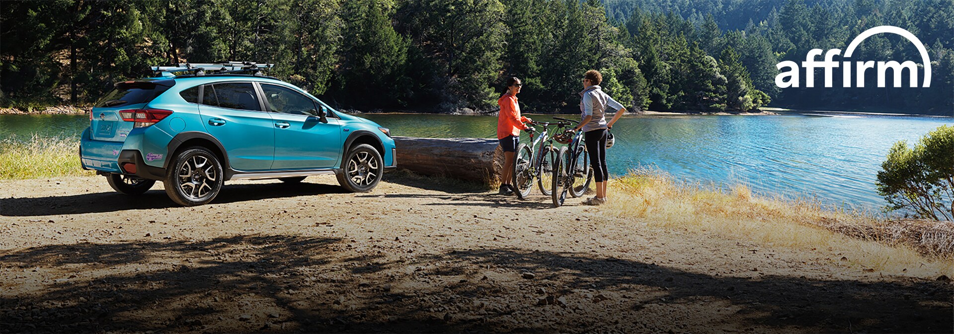 Subaru parked by a lake with roof bike rack. Two people with bikes nearby. Affirm logo.